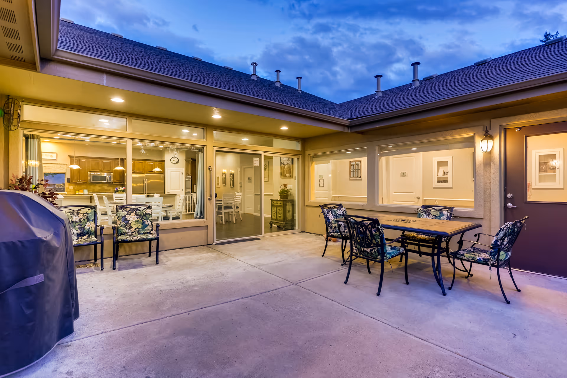 Covered outdoor patio courtyard with a metal dining table and floral-cushioned chairs outside large windows showing the indoor dining/kitchen area.