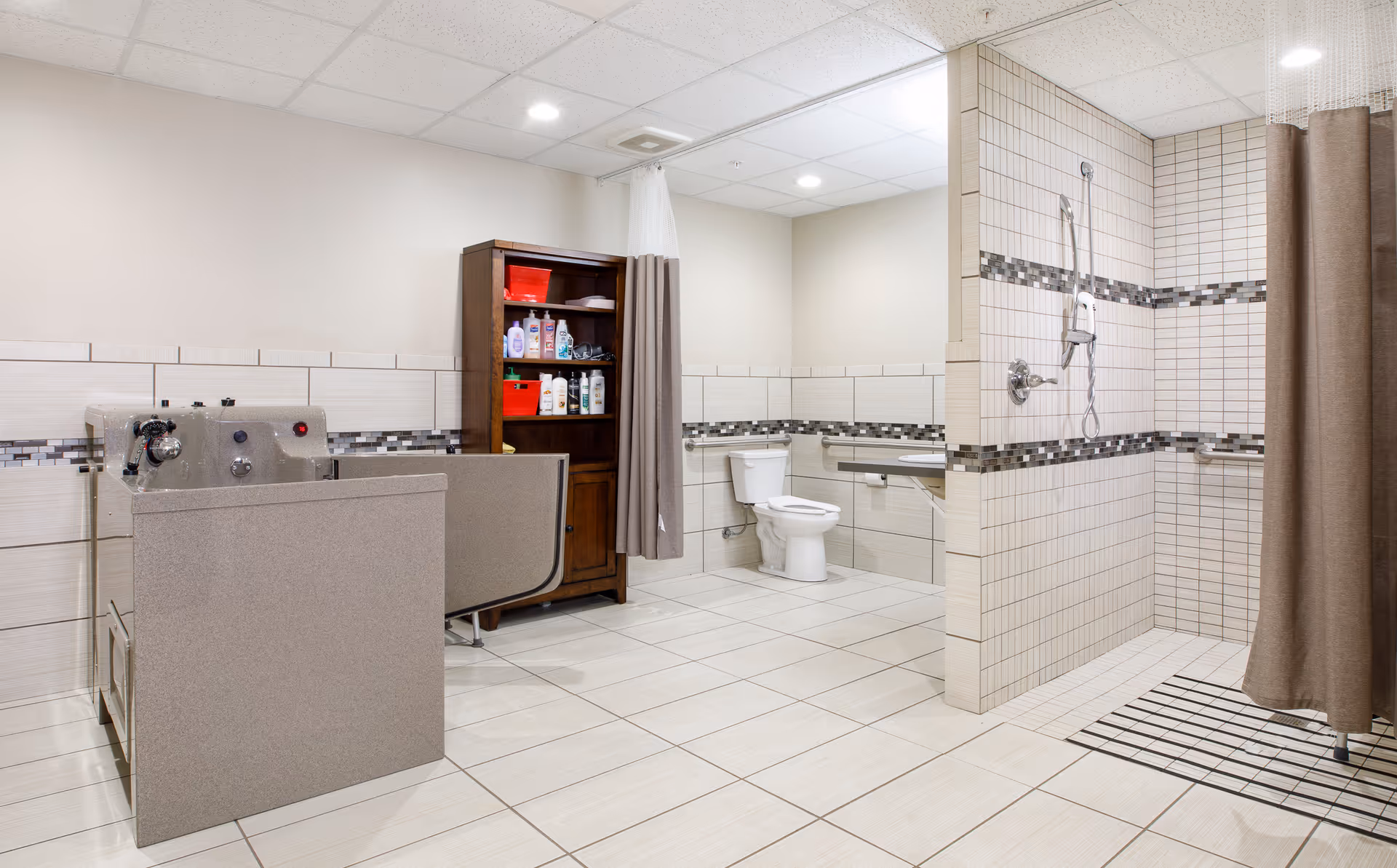 A spacious assisted living bathroom featuring a walk-in shower with grab bars and a handheld showerhead, a toilet with support rails, a wooden cabinet stocked with toiletries, and a bathing tub with privacy curtains. The room has tiled floors and walls with a decorative tile border.