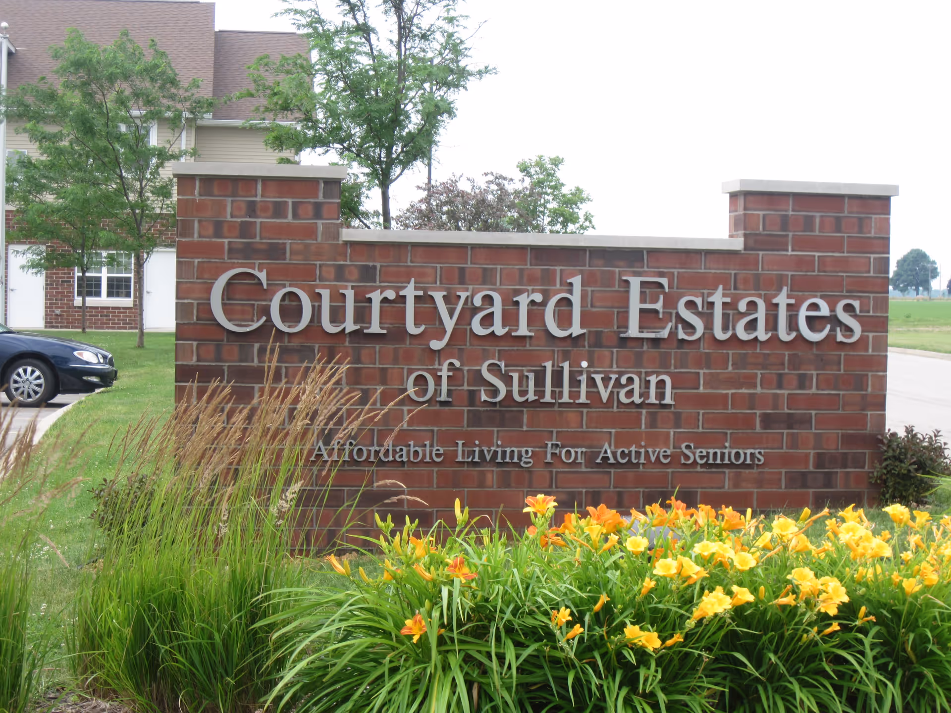 A brick sign for Courtyard Estates of Sullivan with the tagline Affordable Living For Active Seniors, surrounded by green grass, tall plants, and yellow flowers, with a building and a parked car in the background.