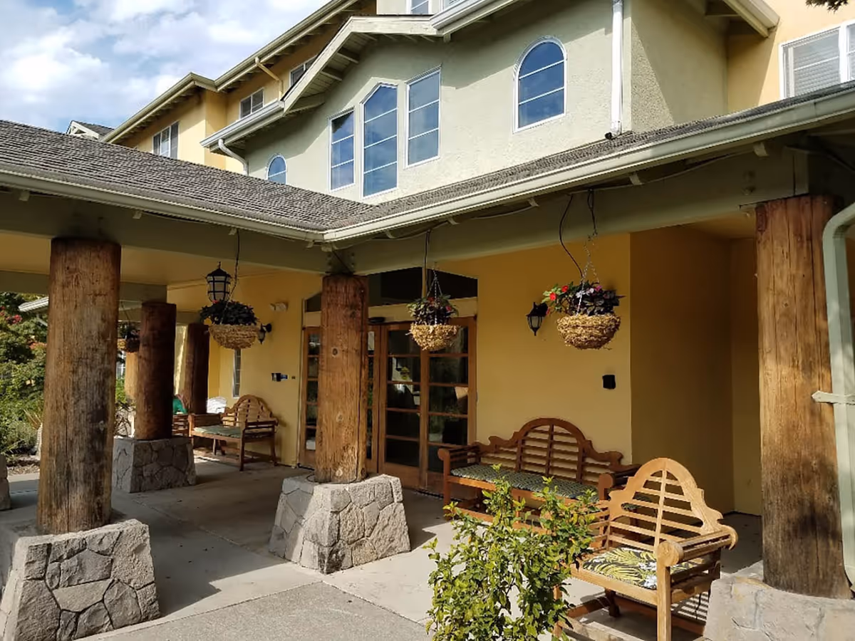 Outdoor covered patio area at Solstice Senior Living at Santa Rosa with wooden benches, hanging flower baskets, large wooden columns with stone bases, and a yellow building exterior with multiple windows.