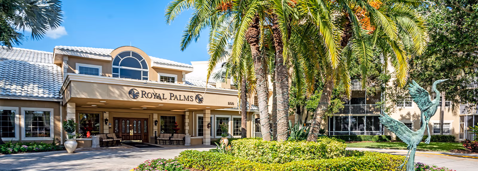 Front entrance of the Royal Palms senior living facility with a covered porte-cochère, palm trees, and a bird sculpture in the landscaped driveway.