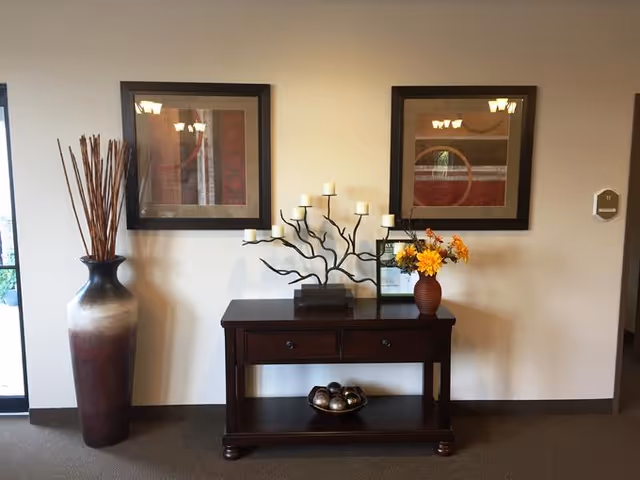 A hallway or lobby area with a dark wooden console table against a beige wall. On the table are a decorative candle holder with multiple white candles, a small vase with yellow and orange flowers, and a framed document. Above the table are two framed abstract art pieces. To the left of the table is a tall vase with long decorative sticks. The floor is carpeted and a door is partially visible on the left side.