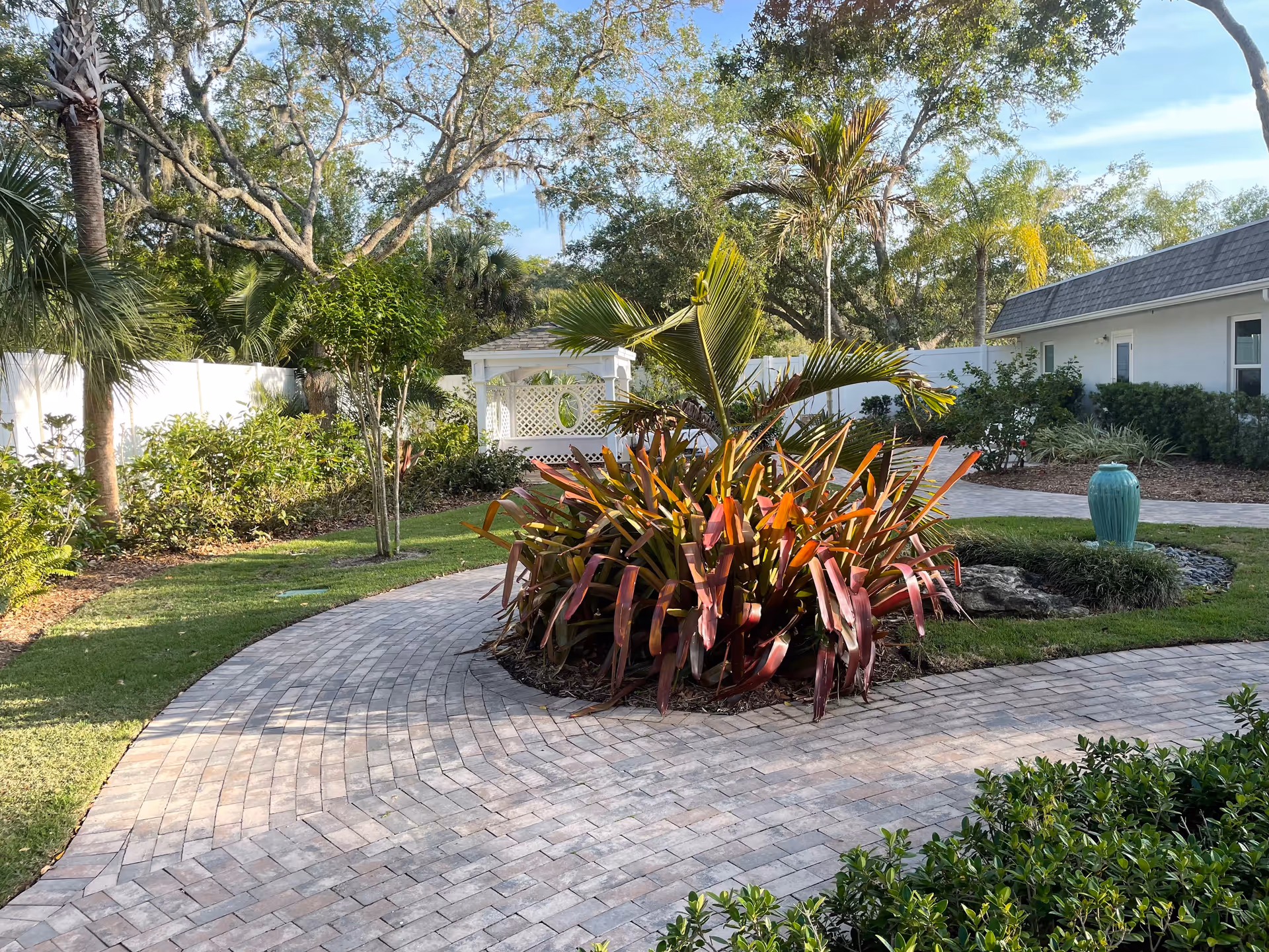 A landscaped outdoor garden area with a curved brick pathway surrounding a large plant with long reddish leaves. There are various trees and shrubs around, a white gazebo in the background, a white fence, and a building with a gray roof on the right side. The sky is clear with some clouds.