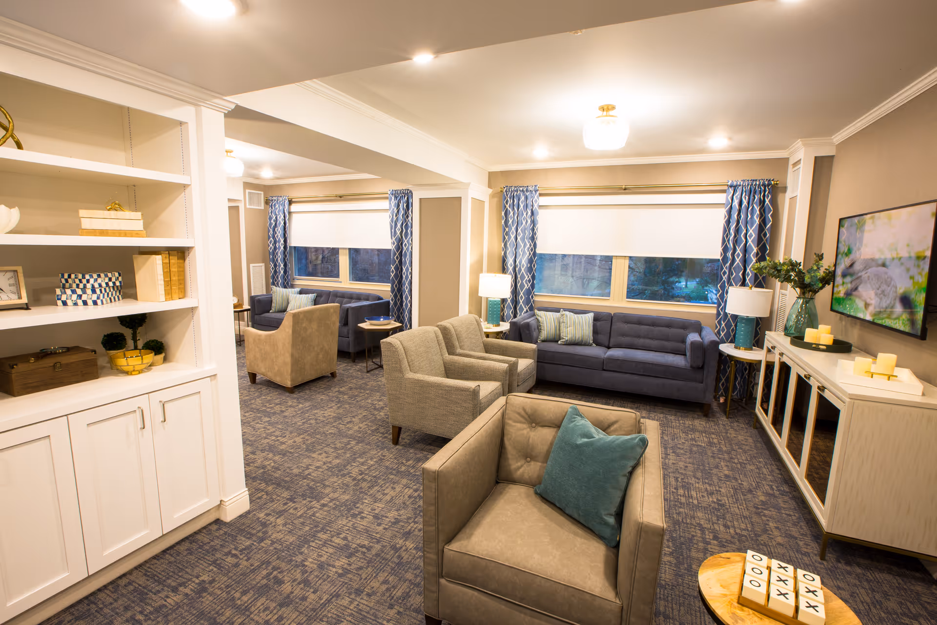A cozy living room area in a senior living facility with multiple armchairs and sofas arranged around small side tables. The room features blue patterned curtains, a carpeted floor, a white built-in shelving unit with decorative items, a wall-mounted TV, and a sideboard with candles and a vase with greenery.