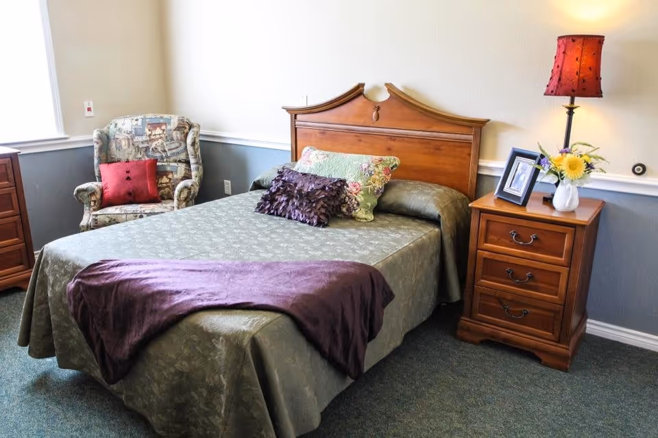 A cozy bedroom with a wooden bed frame, green bedspread, and decorative pillows. There is a wooden nightstand with a red lamp, a framed photo, and a vase with flowers. A patterned armchair with a red cushion is placed near a window with white blinds.