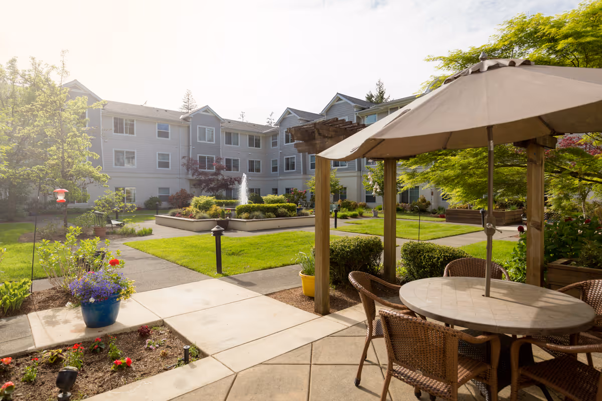 Outdoor courtyard area of a senior living facility with a round table and four wicker chairs under a large beige umbrella. The courtyard features green lawns, a central fountain, flower beds, and a multi-story residential building in the background under a bright sky.