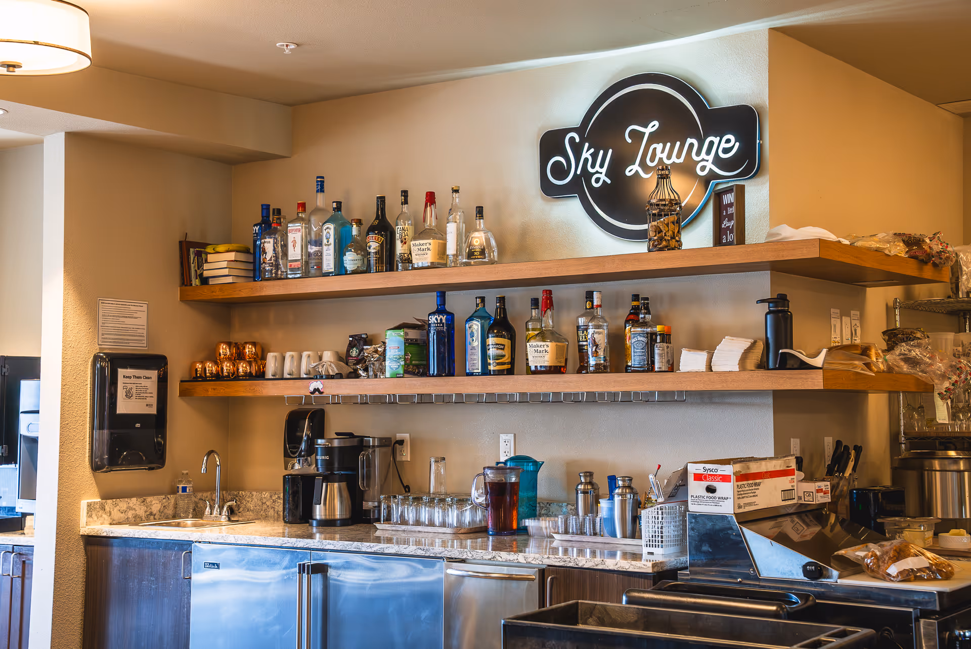 Interior view of a lounge area with a countertop and shelves stocked with various bottles of liquor, coffee makers, glasses, and other drinkware. A neon sign on the wall reads 'Sky Lounge'. The countertop has a sink and some kitchen supplies, with a paper towel dispenser mounted on the wall.