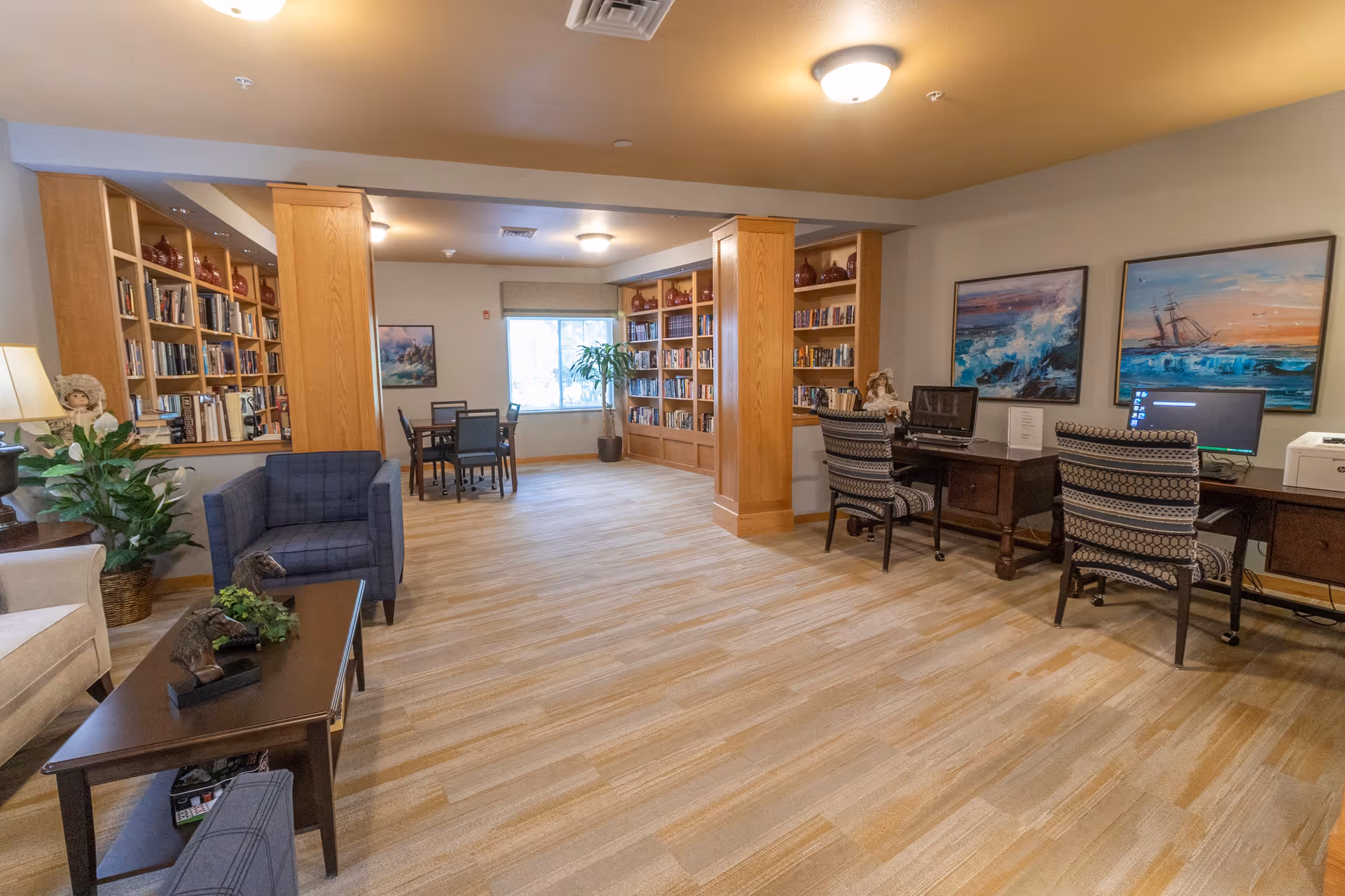 A spacious and well-lit common area in a senior living facility featuring wooden bookshelves filled with books, a seating area with armchairs and a coffee table, two desks with computers and chairs, and framed paintings on the wall. There is a window at the back letting in natural light and a potted plant near the window.