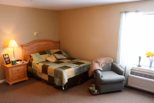 A cozy bedroom with a wooden bed frame and a patchwork quilt on the bed. There is a wooden nightstand with a lamp, a framed photo, and some items on it. Next to the bed is a comfortable armchair with a blanket draped over one arm. A window with sheer curtains allows natural light into the room, and below the window is a heating or air conditioning unit with a vase of flowers and a candle on top.