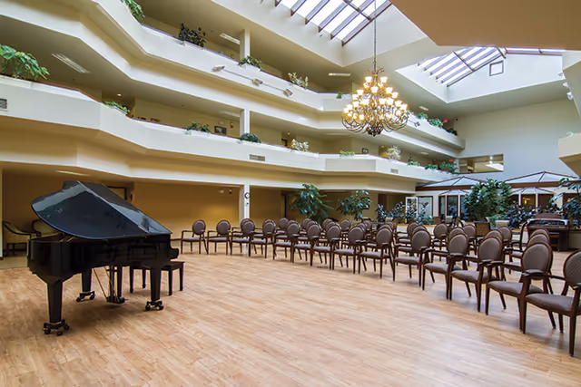 Spacious indoor common area with a grand piano on the left and several rows of chairs arranged facing the piano. The room has a high ceiling with skylights and a large chandelier hanging in the center. Multiple floors with balconies and plants overlook the area.