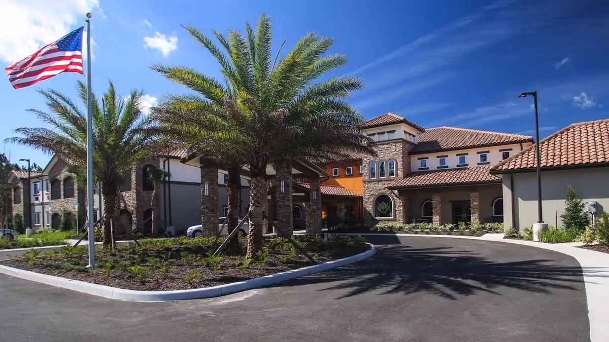Mediterranean-style senior living building entrance with palm trees, an American flag, and a curved driveway under a blue sky.