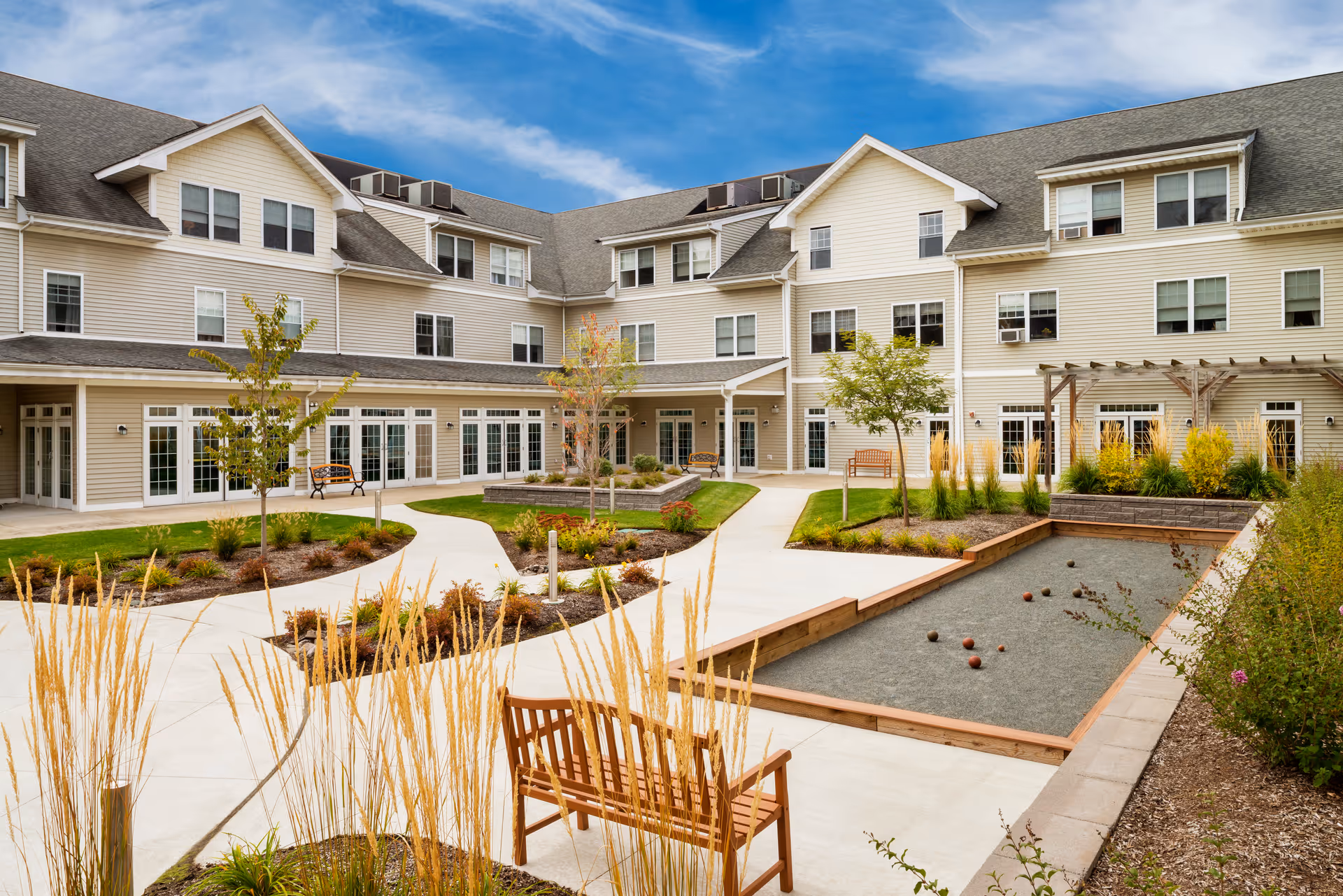 Outdoor courtyard area of a senior living facility with paved walkways, landscaped garden beds, benches, small trees, and a bocce ball court, surrounded by a three-story beige building under a partly cloudy sky.
