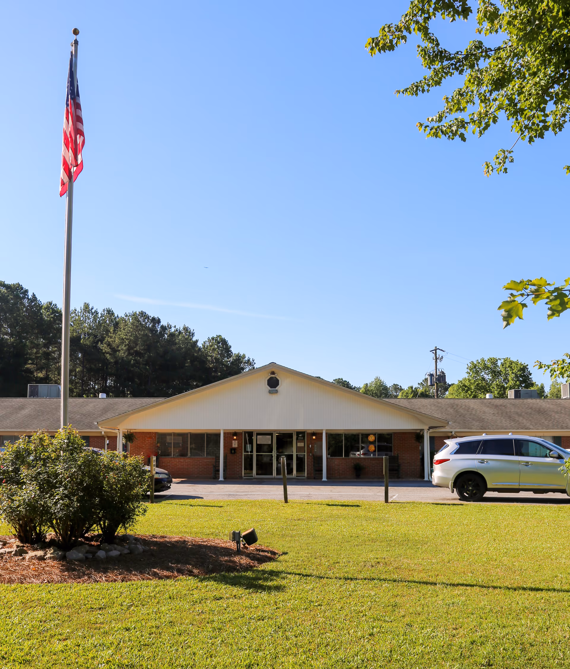 Front exterior view of Greene Point Health & Rehabilitation building with a flagpole displaying the American flag, a green lawn, bushes, and a parked silver SUV under a clear blue sky.