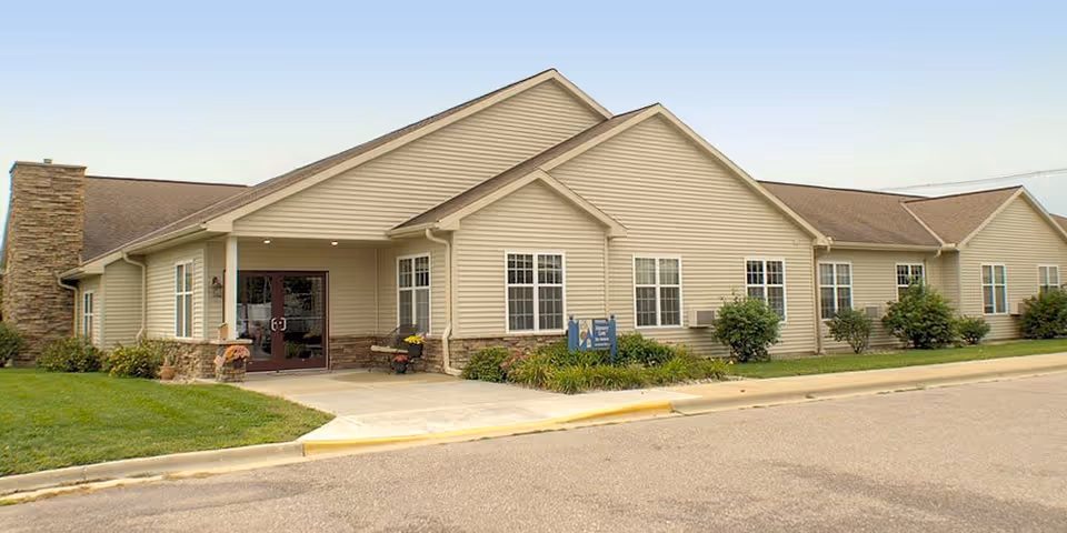 Exterior view of a single-story beige senior living facility building with a peaked roof, multiple windows, a stone chimney on the left side, and a covered entrance with double glass doors. There is a small landscaped area with bushes and a sign near the entrance, and a paved driveway in front.