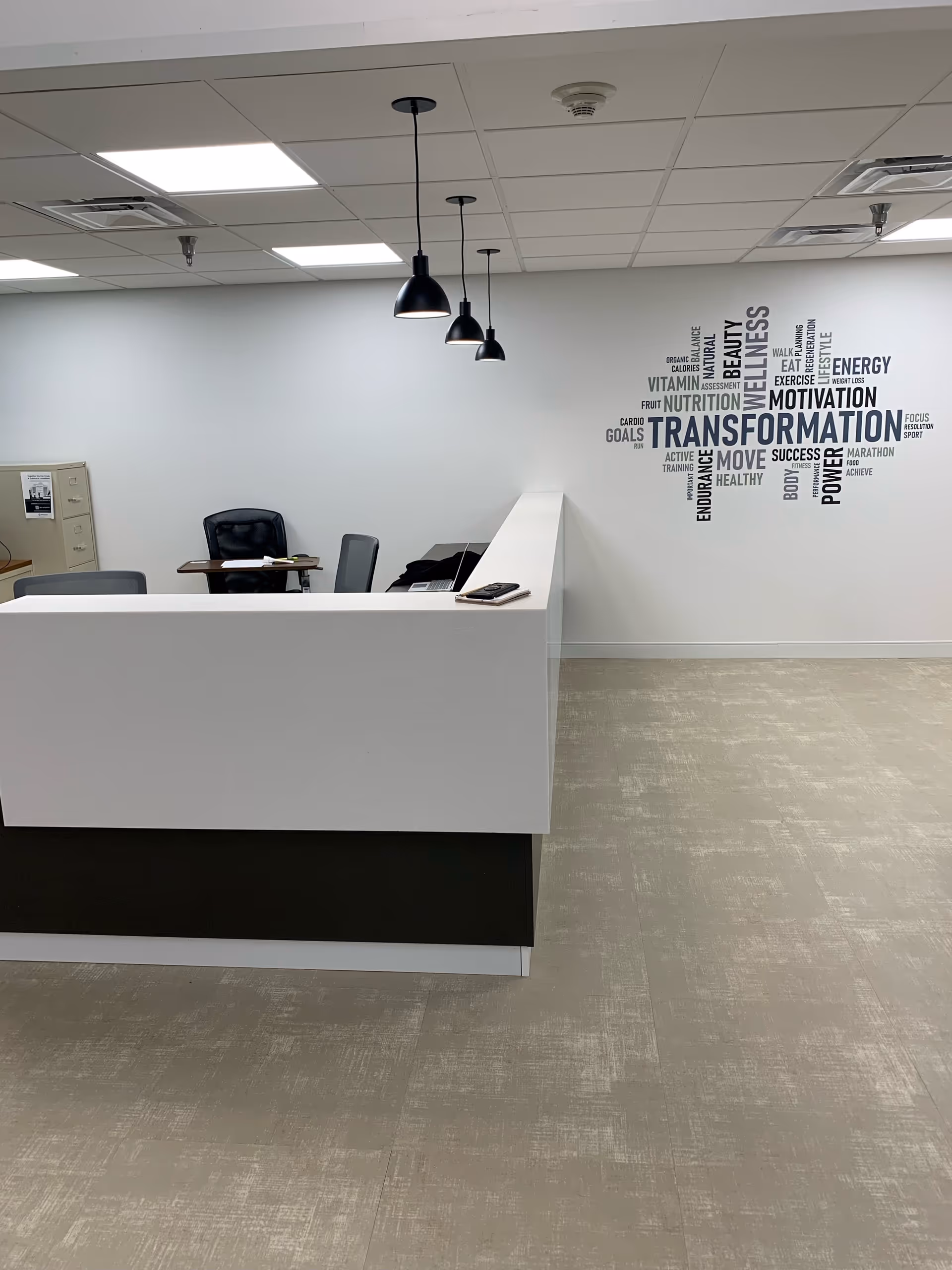 Reception area with a white and black desk, three black pendant lights hanging from the ceiling, office chairs and desks in the background, and a wall decal with words related to wellness and transformation.