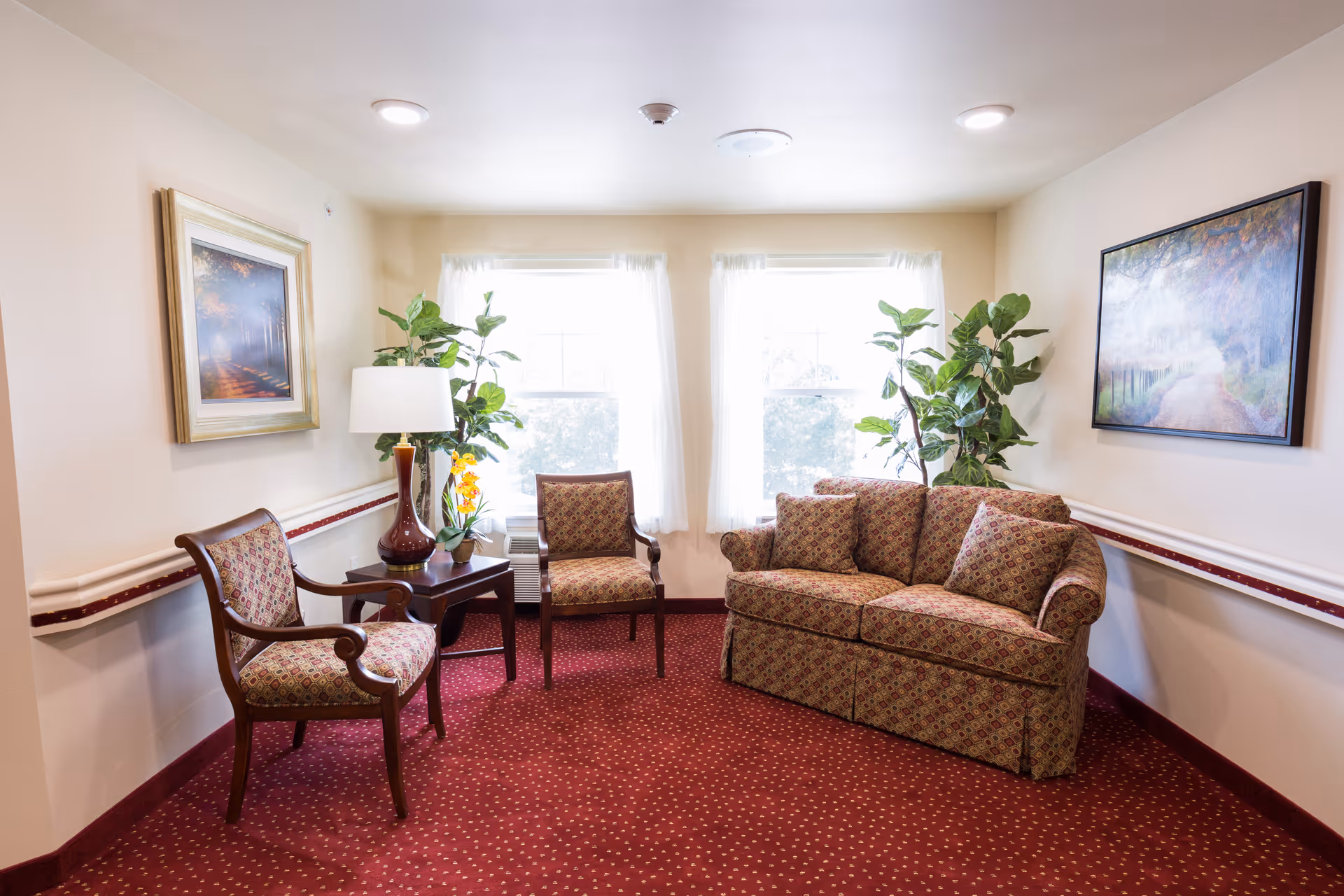 Bright sitting area with a patterned sofa and chairs, a small table with a lamp and plants in front of two windows.