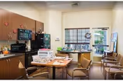 Interior view of a small kitchen and dining area in an assisted living facility. The room features wooden cabinets, a black microwave, stove, and refrigerator. A table with chairs is in the center, with some papers and containers on it. There is a window with blinds and a glass door leading outside. Additional chairs are arranged along the wall.