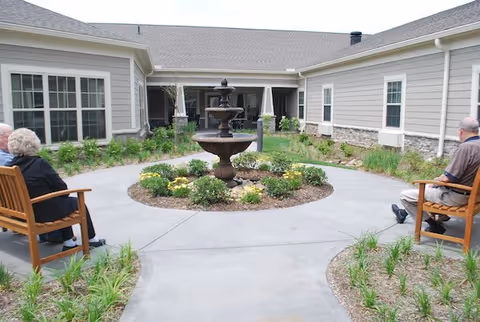 Sunlit courtyard with a three-tier fountain at the center, planted beds, paved walkways, benches and a surrounding single-story building.
