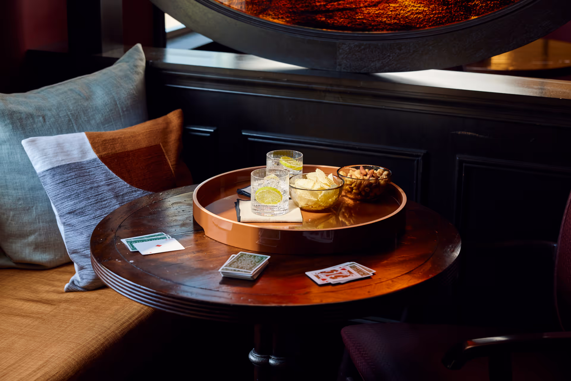 Round wooden table holding a tray with drinks, bowls of chips and nuts, and scattered playing cards next to a cushioned seat.