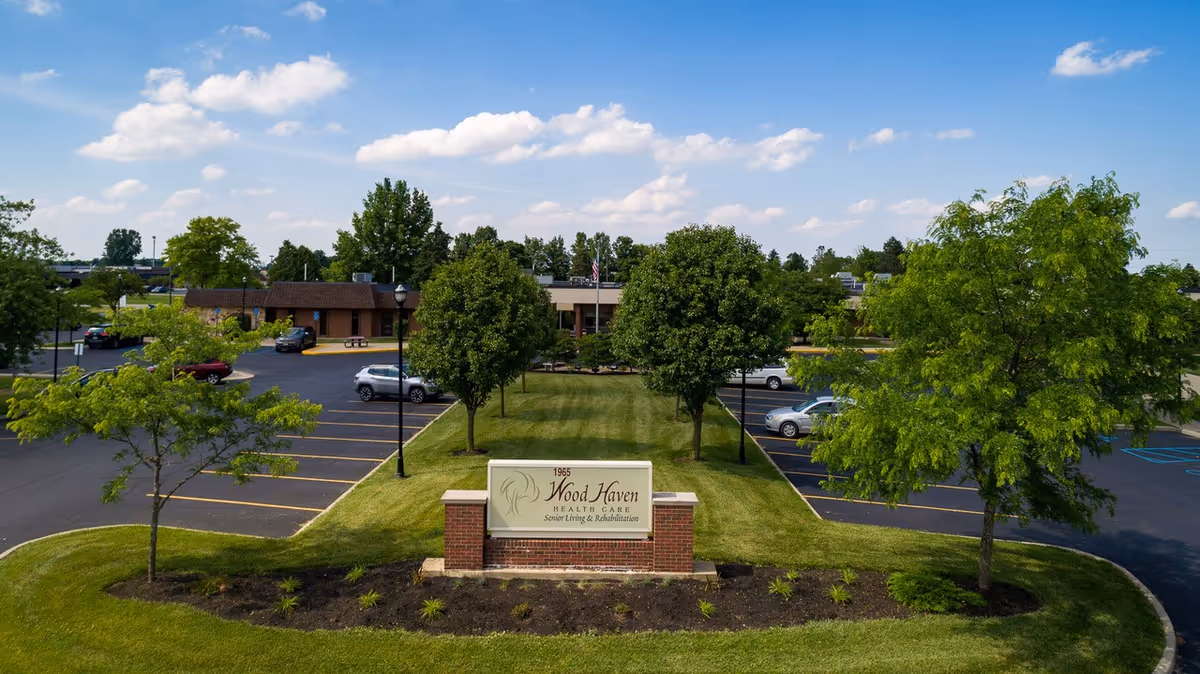 Exterior view of Wood Haven Healthcare facility showing a parking lot with several cars, a grassy area with trees, and a sign in the foreground that reads 'Wood Haven Health Care Senior Living & Rehabilitation'. The sky is partly cloudy.
