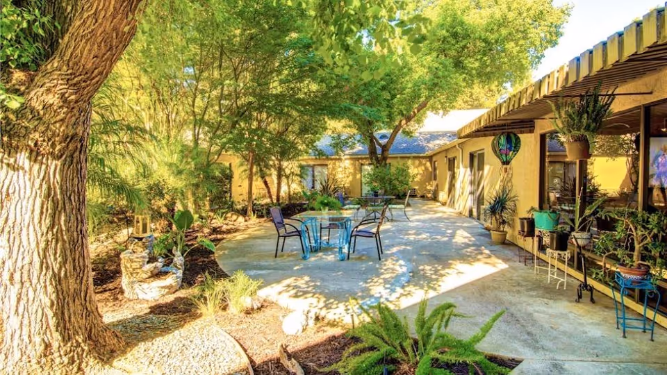 Outdoor patio area at Greenhaven Estates Assisted Living and Memory Care featuring a concrete walkway with a glass table and four chairs. The area is surrounded by lush green trees, plants, and garden decorations, with a building wall on the right side adorned with hanging plants and a colorful hot air balloon decoration.
