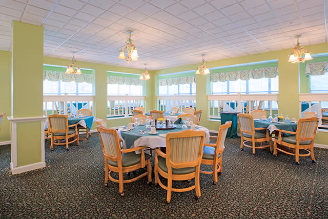 A bright dining room with multiple round tables covered with green tablecloths and set with glasses, plates, and napkins. The room has large windows with floral valances, light green walls, and hanging light fixtures. Wooden chairs with green cushions surround the tables.