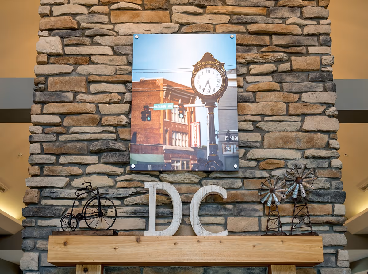A stone wall with a wooden mantel shelf displaying decorative items including a small metal bicycle sculpture, large white letters 'DC', and two metal windmill sculptures. Above the mantel is a framed photo of a street clock with the words 'City of Greenwood' and a street sign for Madison Ave.