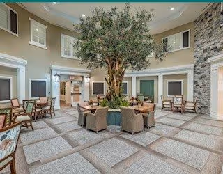 Spacious indoor atrium lounge with a large central indoor tree surrounded by chairs and tables beneath a high ceiling.