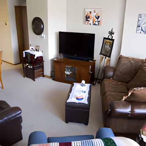 A cozy living room with a brown leather sofa and armchair, a flat-screen TV on a wooden stand, a small table with a white cloth and decorative items, and a carpeted floor. The walls have framed artwork and a decorative mirror.
