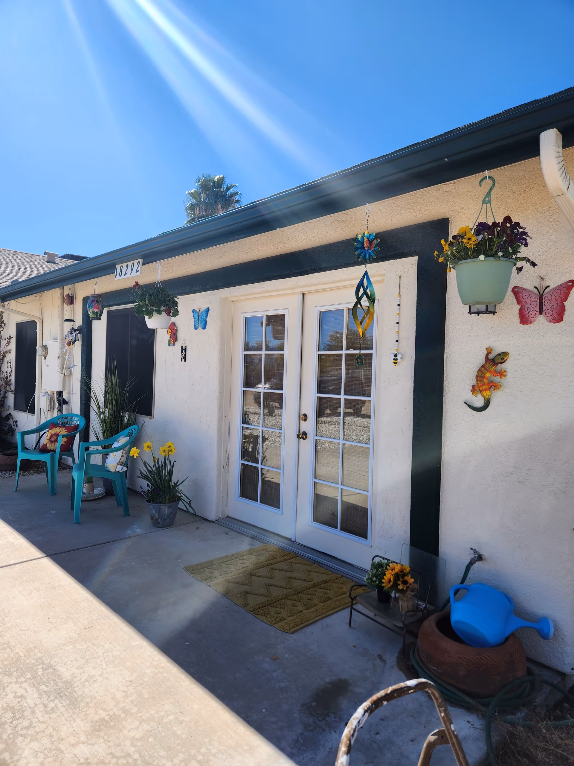 Exterior view of a single-story building with white walls and a dark green trim. There are two turquoise chairs with cushions, potted plants, and hanging flower pots decorating the entrance area. The entrance features white double French doors with glass panes. Various colorful decorations, including butterflies and a lizard, adorn the wall. The building number 18292 is displayed above the door. The sky is clear and blue with sunlight streaming down.
