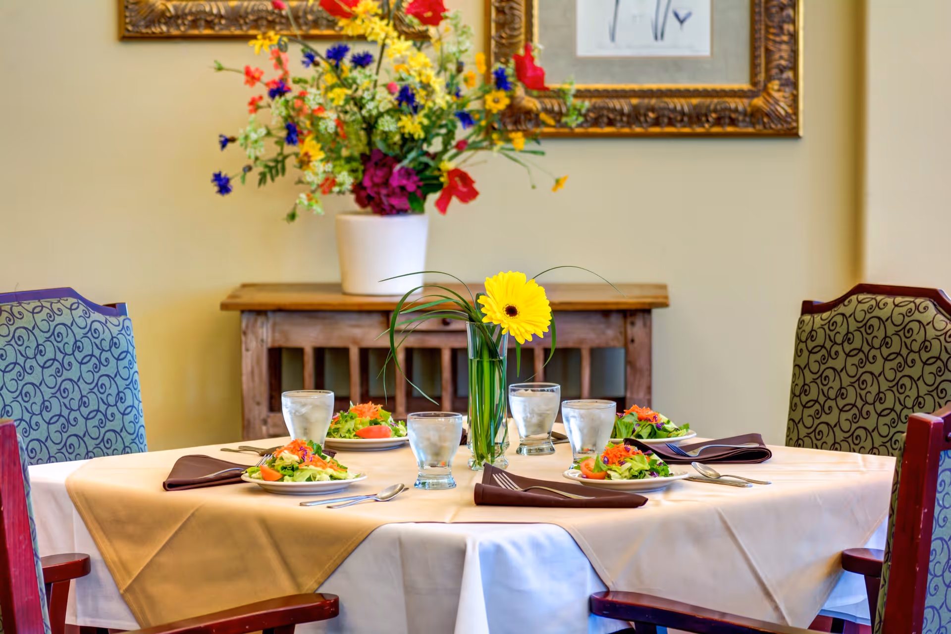 A dining table set for four with plates of salad, glasses of water, silverware, and brown napkins. A vase with a yellow flower and green leaves is in the center of the table. Behind the table is a wooden sideboard with a large colorful flower arrangement and framed artwork on the wall.