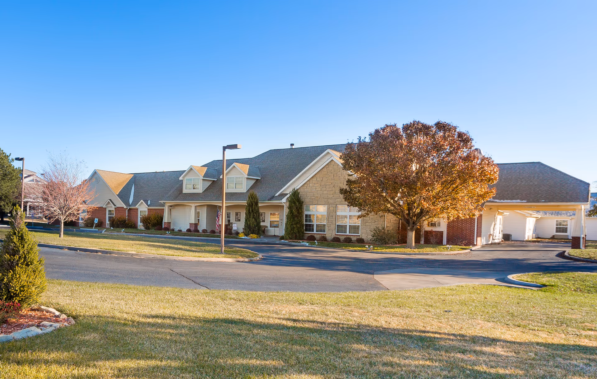 Exterior view of a single-story senior living facility building with a combination of brick and stone facade, surrounded by a well-maintained lawn and trees with autumn foliage under a clear blue sky.