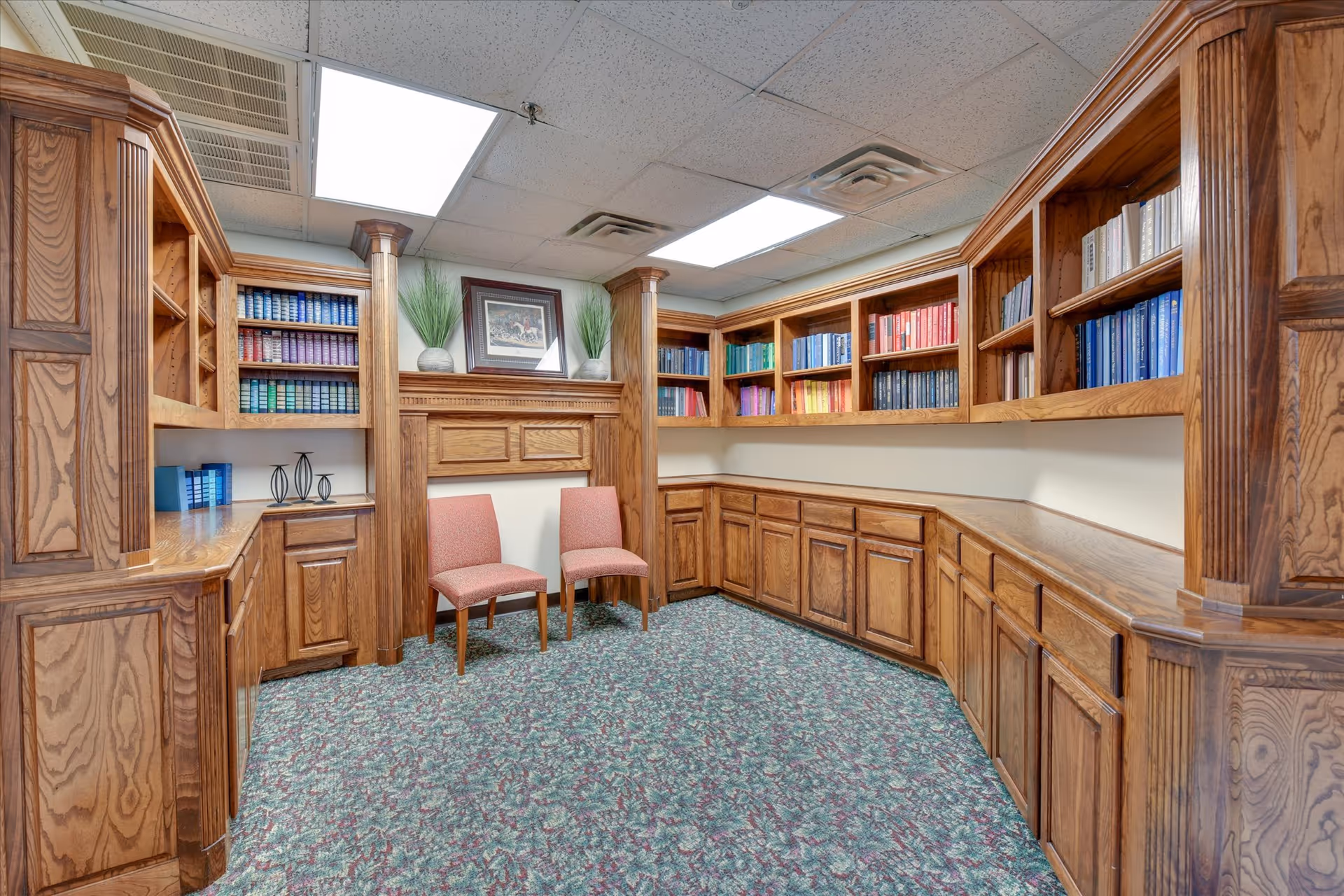 A cozy library or reading room with wooden built-in bookshelves filled with colorful books, two pink upholstered chairs, a framed picture, and decorative plants on a wooden mantel. The room has a patterned carpet and fluorescent ceiling lights.