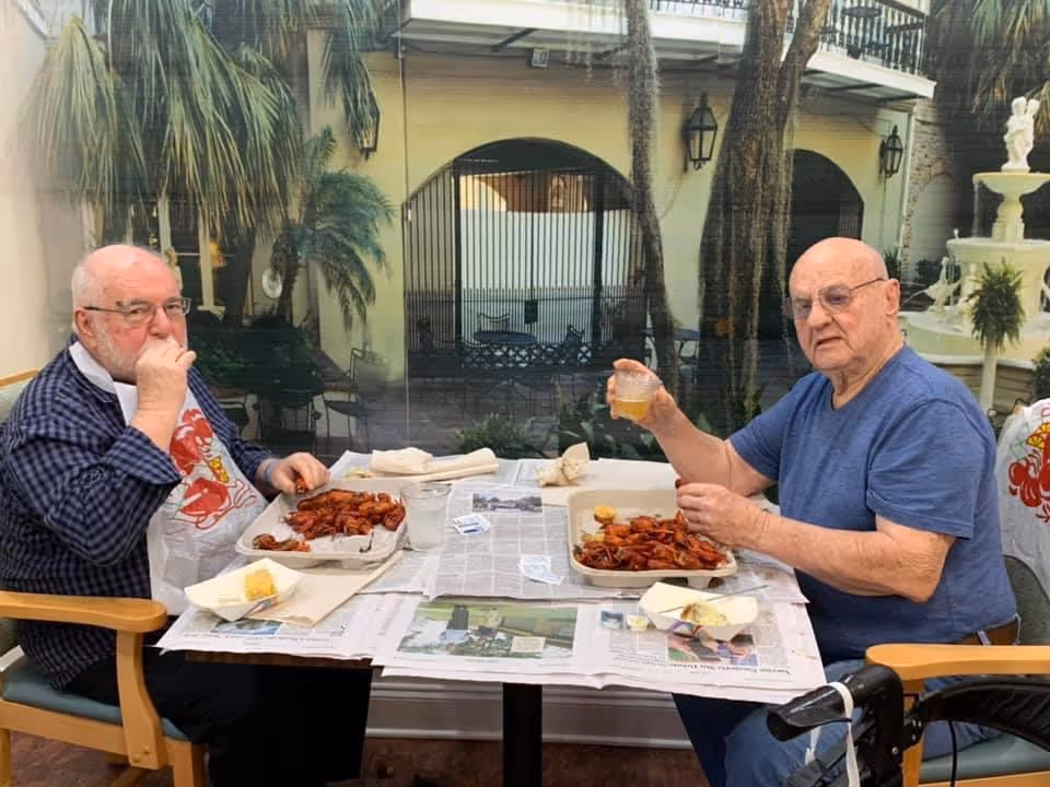 Two elderly men sitting at a table covered with newspapers, eating a meal of crawfish. One man is wearing a bib and a checkered shirt, while the other is in a blue t-shirt holding a drink. They are seated indoors near a large window showing an outdoor courtyard with palm trees and a fountain.