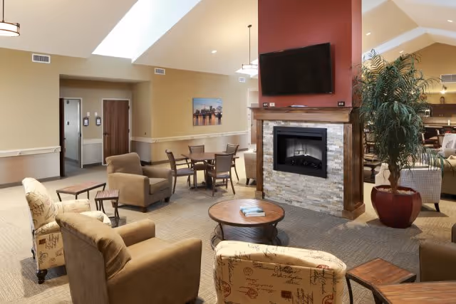 A spacious living room area with multiple armchairs and small tables arranged around a central stone fireplace with a mounted flat-screen TV above it. The room has high ceilings with skylights, beige walls, and a large potted plant near the fireplace. There is a dining table with chairs in the background and a doorway leading to another room.