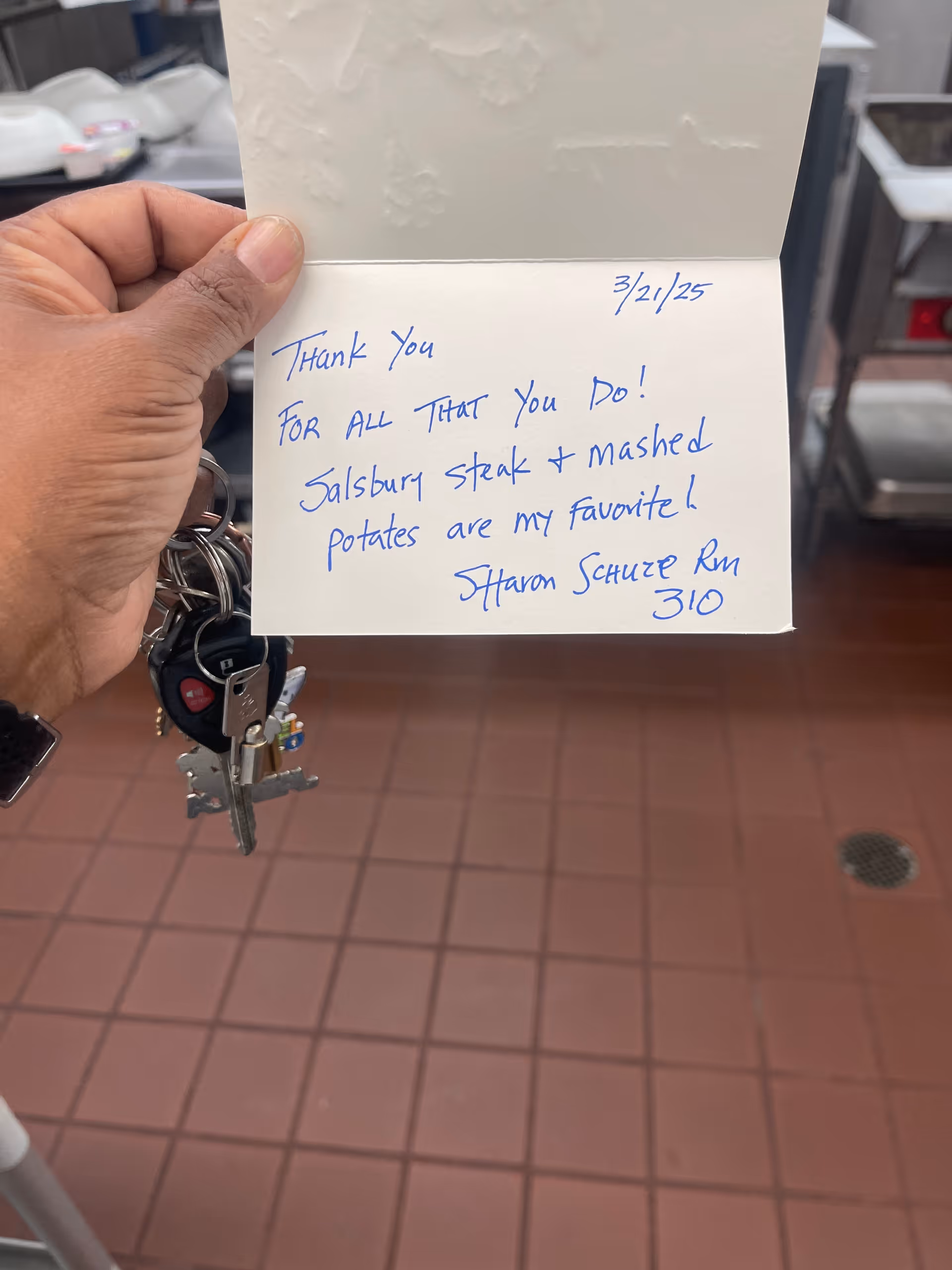 A hand holding a handwritten thank-you card and a set of keys over a tiled commercial kitchen floor with stainless equipment in the background.