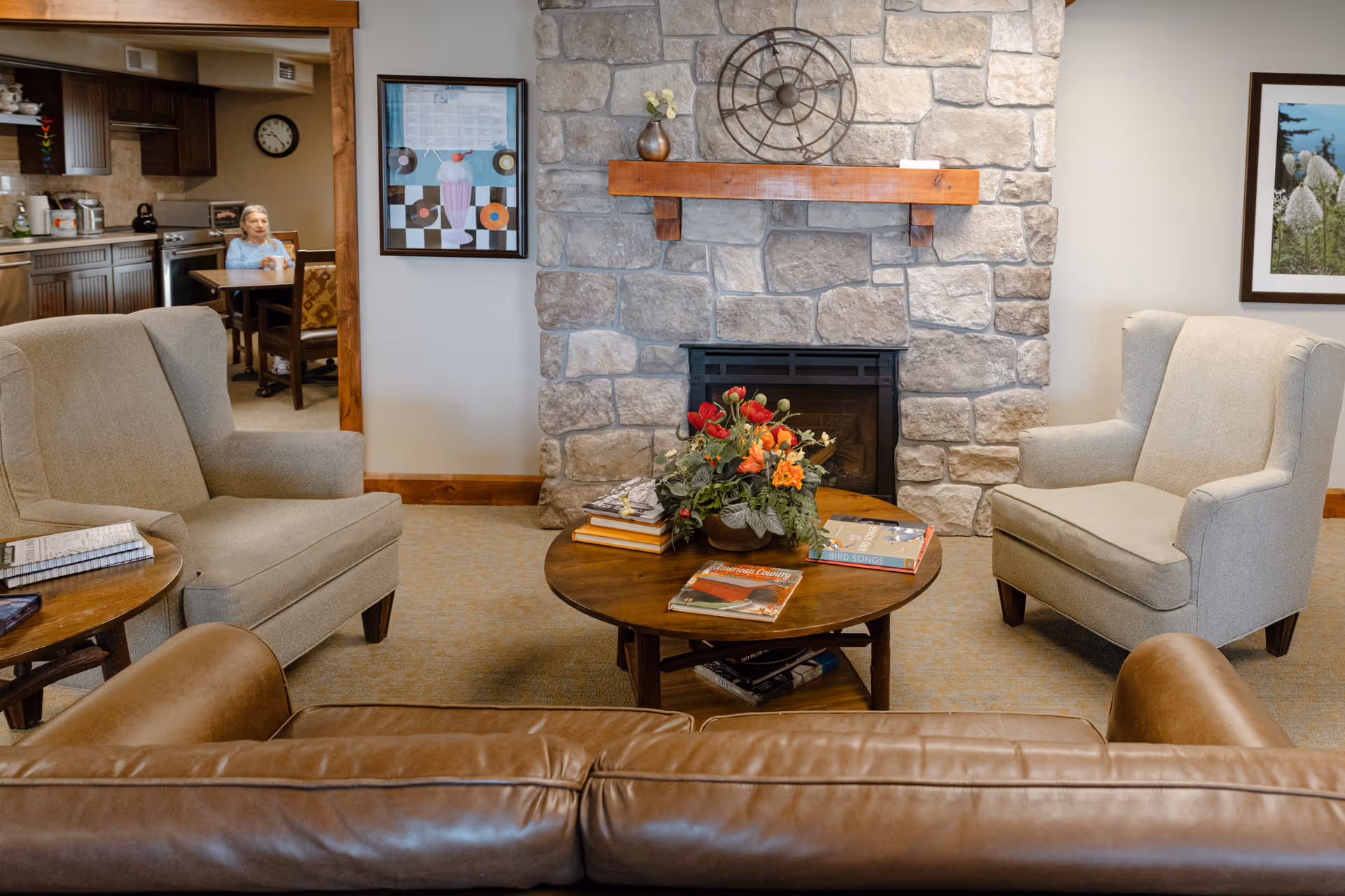 A cozy living room area with a stone fireplace in the center, flanked by two beige armchairs. In front of the fireplace is a round wooden coffee table with a floral arrangement and several books on it. A brown leather sofa is partially visible in the foreground. In the background, there is a dining area with a woman sitting at a table. The walls are decorated with framed artwork.
