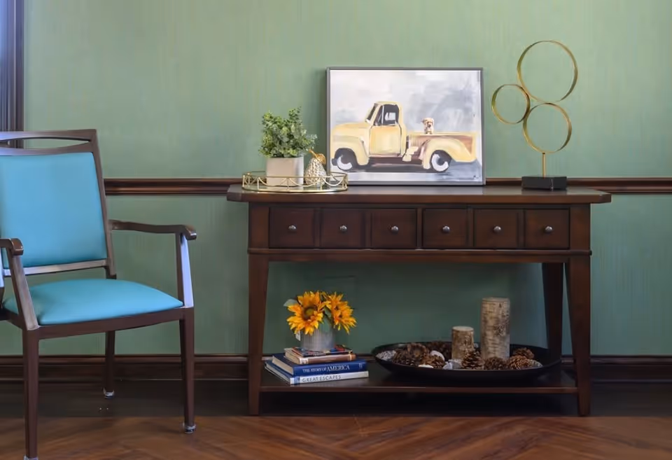 A wooden console table with six drawers against a green wall with wood trim. On top of the table is a small potted plant, a decorative gold pineapple, a tray, a painting of a yellow pickup truck with a dog in the bed, and a modern gold sculpture with three circular rings. Below the table are two books, a vase with sunflowers, and a black tray filled with pinecones and decorative logs. To the left of the table is a wooden chair with turquoise upholstery.