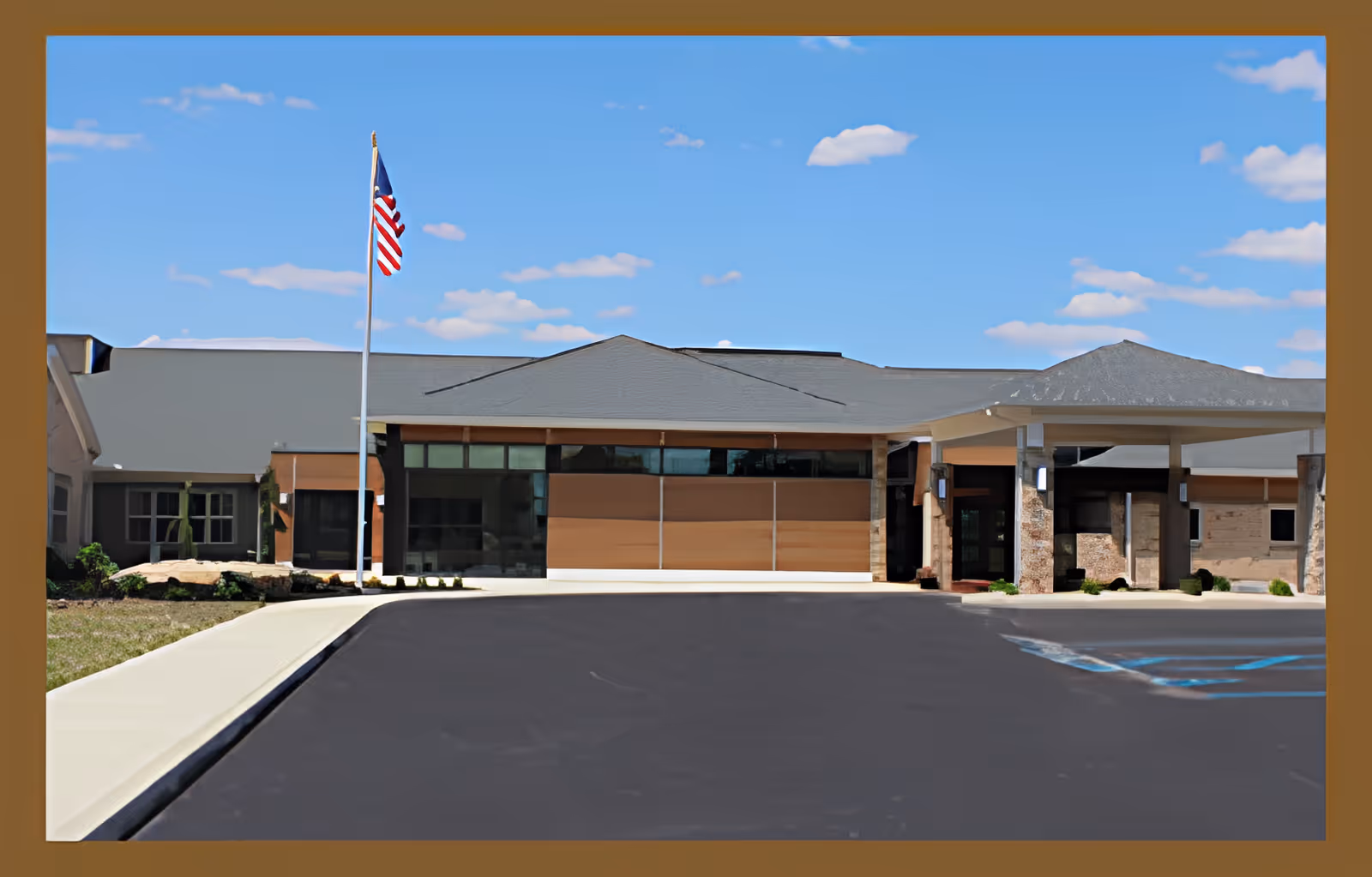 Front entrance of a single-story senior living facility with an American flag, covered drop-off, and an empty parking area under a blue sky.