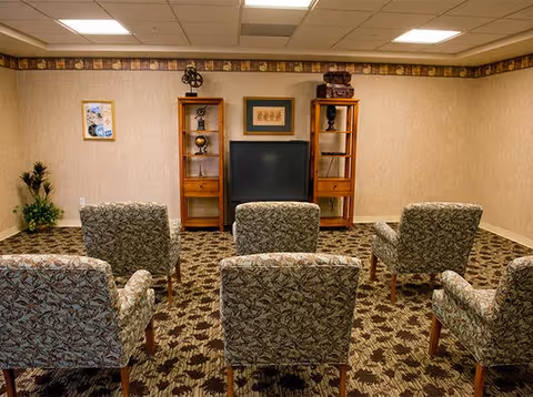 A small common room with two rows of upholstered patterned armchairs facing a television flanked by wooden shelving units and framed wall art.