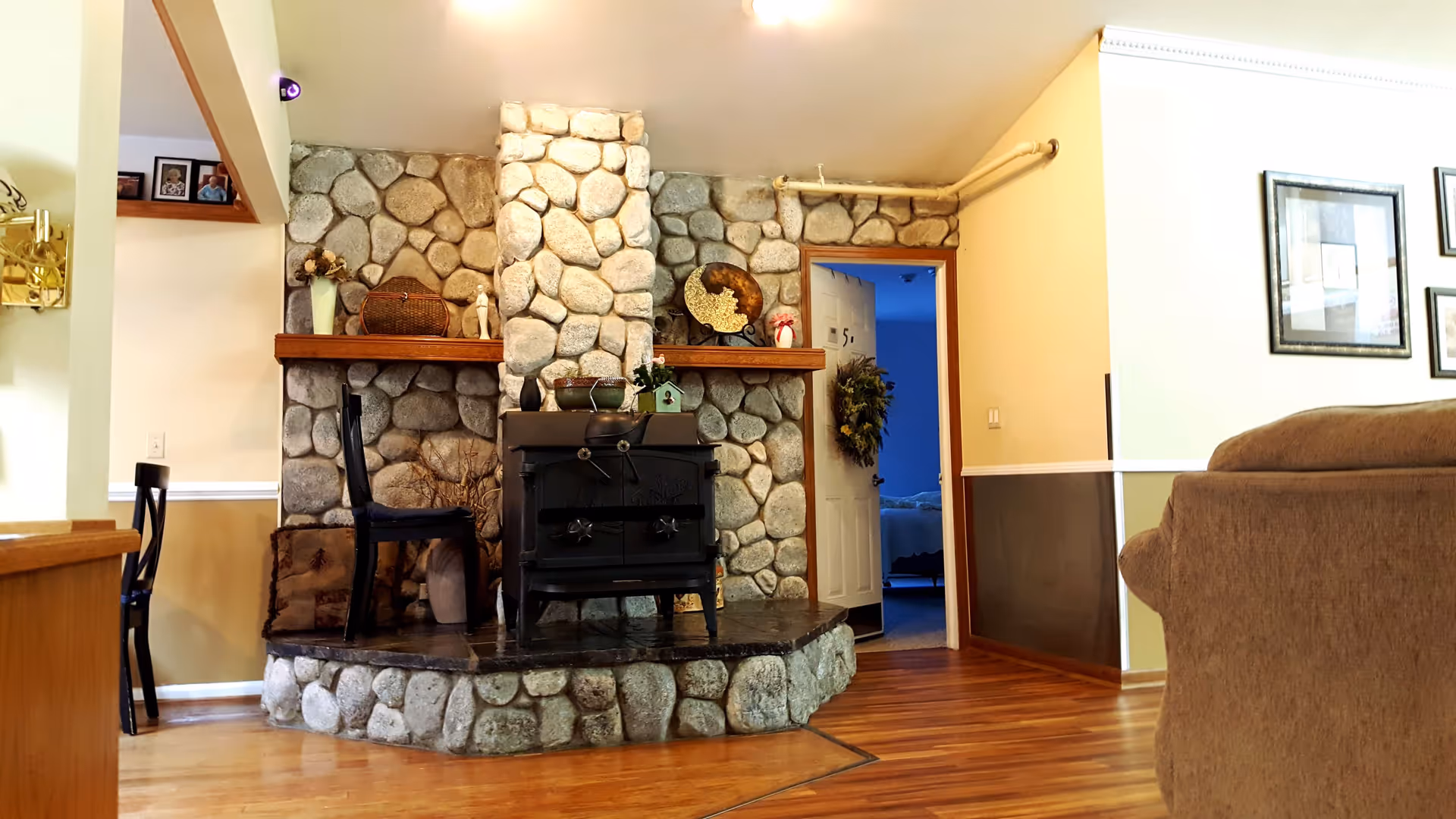 Interior view of a cozy living room area featuring a stone fireplace with a black wood stove. There are decorative items on the wooden mantel, a chair placed next to the fireplace, and a partially open door leading to a room with a bed. The room has wooden flooring and framed pictures on the wall.