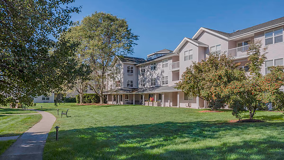 A large, multi-story residential building with balconies and many windows, surrounded by green grass, trees, and a paved walkway under a clear blue sky.