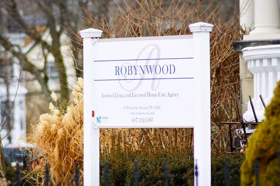 White post sign for Robynwood Assisted Living and Licensed Home Care Agency standing among shrubs in front of a building column.