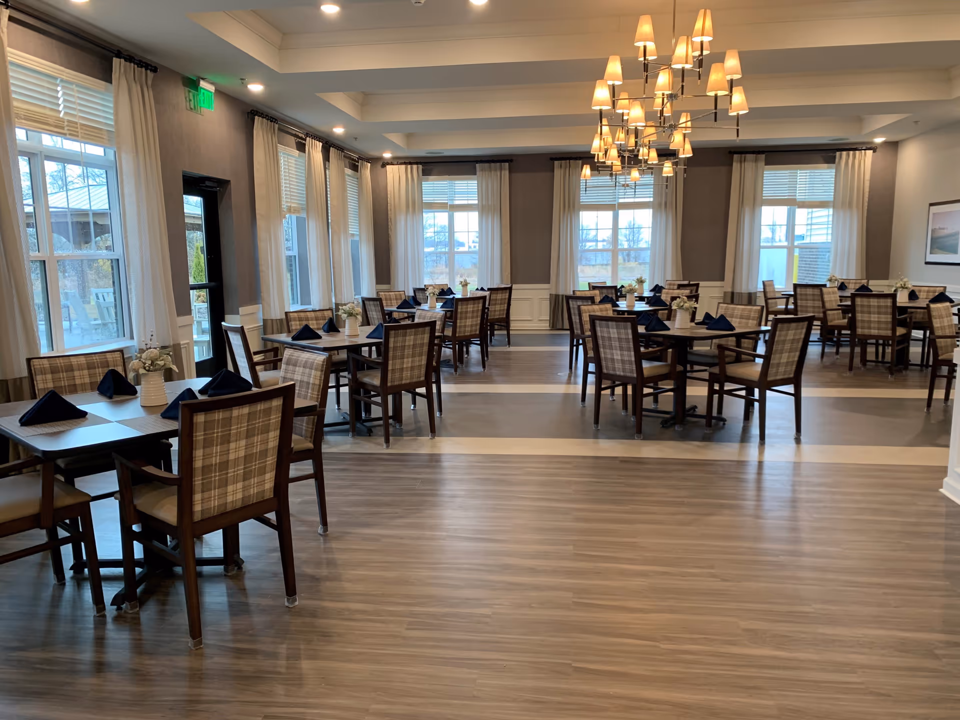 A spacious dining room with multiple wooden tables and plaid cushioned chairs. Each table is set with dark blue folded napkins and small white flower arrangements. Large windows with white curtains allow natural light to fill the room. A modern chandelier with multiple lampshades hangs from the ceiling.