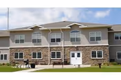 A two-story senior living building with stone facade, windows, door, flagpole, and grassy frontage.