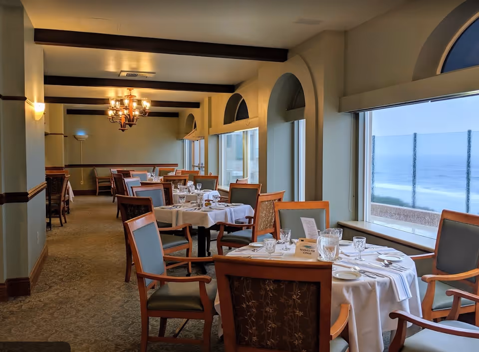 Dining room with neatly set tables and chairs and large arched windows looking out to the ocean.