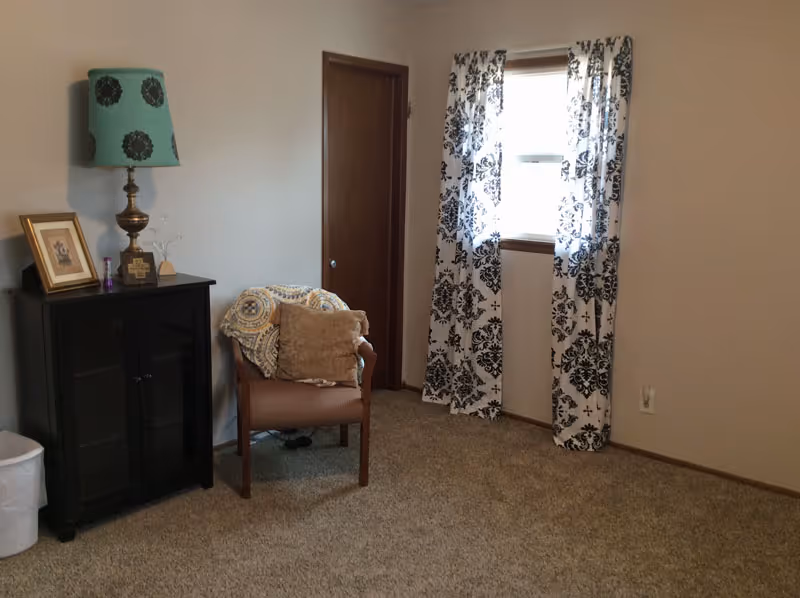 A cozy room with beige carpet and light-colored walls. There is a wooden chair with a cushion and a patterned throw blanket draped over it. Next to the chair is a small black cabinet with a decorative lamp, a framed picture, and a small cross on top. A window with white and black patterned curtains lets in natural light. A closed wooden door is visible in the background.