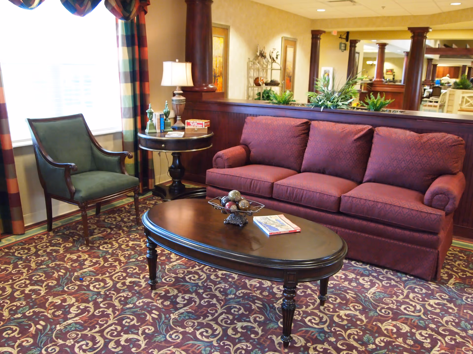 A cozy living room area with a maroon patterned sofa, a dark wooden oval coffee table with decorative items and a book on it, a green upholstered armchair, a round side table with a lamp and decorative bottles, and colorful plaid curtains on the window. The room has a patterned carpet and wooden columns with plants on a divider behind the sofa.