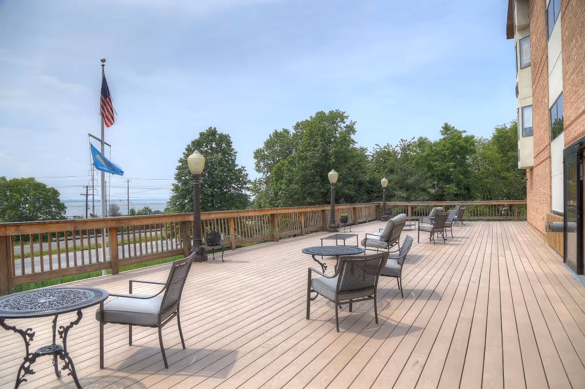 Spacious wooden outdoor deck with seating, lamps, and a flag beside a brick building.