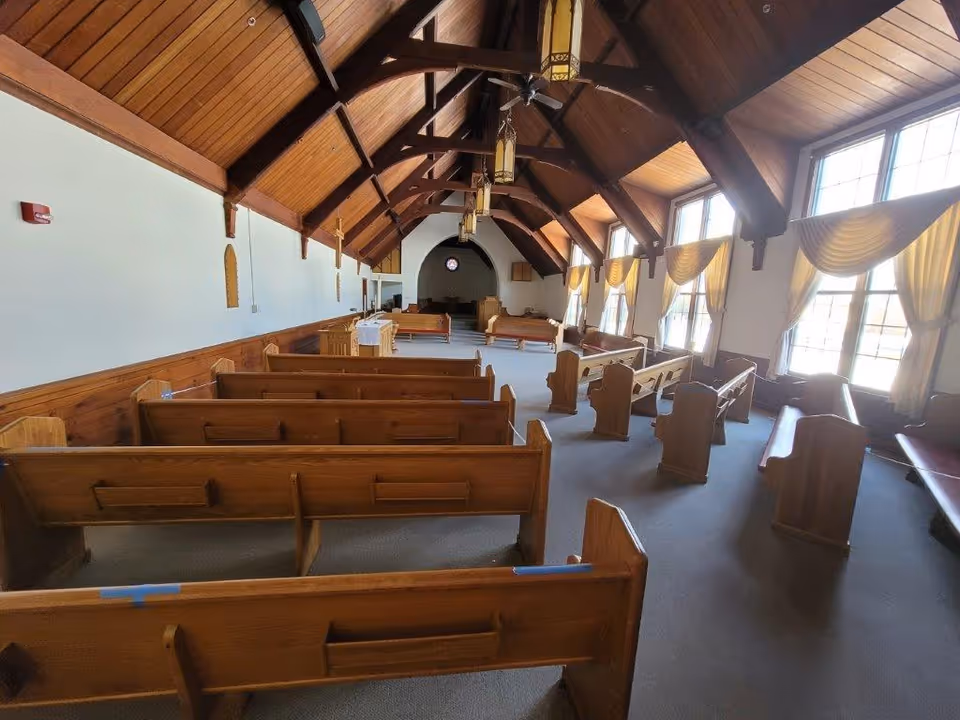 Interior of a chapel or worship room with wooden pews arranged in rows, large windows with yellow curtains letting in natural light, and a high wooden vaulted ceiling with hanging lantern-style lights.